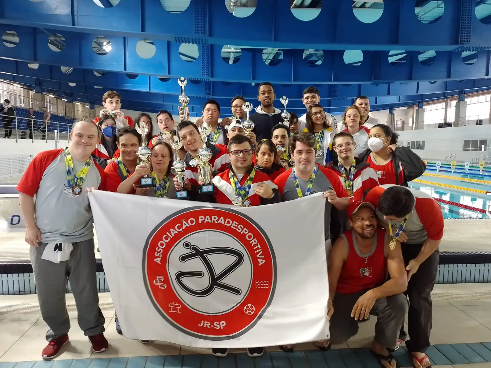 Pessoas reunidas atrás da bandeira da Associacao Paradesportiva JR Natacao