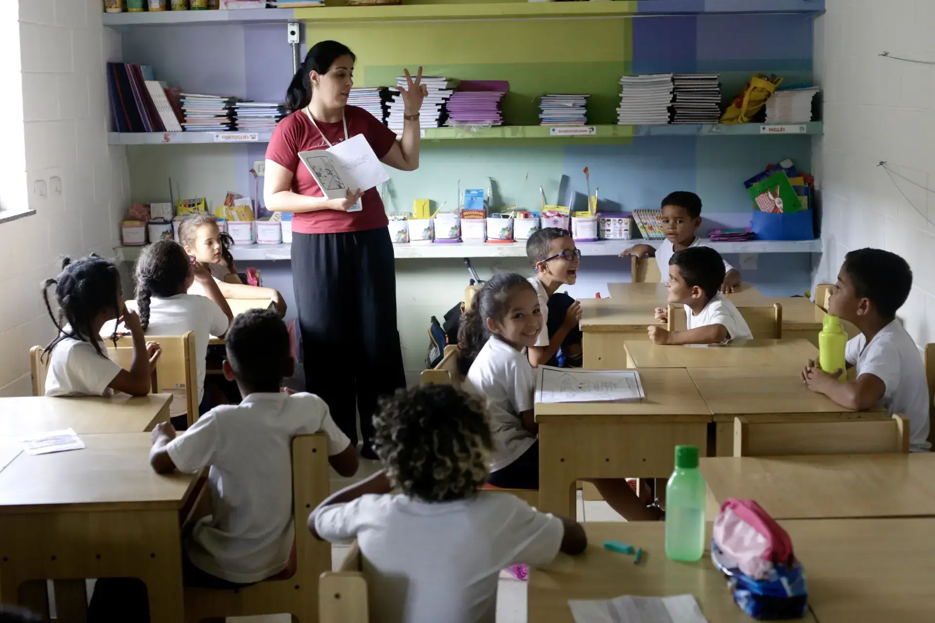 Crianças ouvindo estórias em uma sala de aula por uma professora em pé. Projeto Mão Amiga